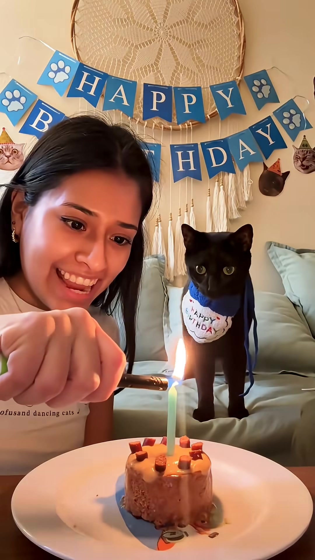 The owner smiles as she lights a candle on the cat's birthday cake, with the cat looking on.