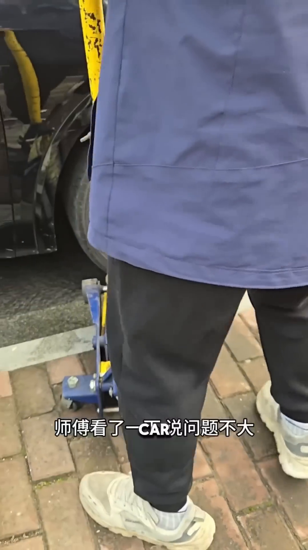 A person is lying on the ground under a car, holding a small kitten. Another person is operating a car jack.